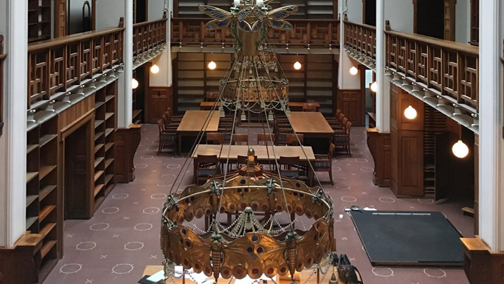 Et kig ned i det tidligere bibliotek fra 1. sals højde. I forgrunden ses den store lysekrone. Reoler samt interiør er i mørkt træ, mens høje, hvis søjler rejser sig mod loftet. Der er læseborde med stole bagerst i salen.