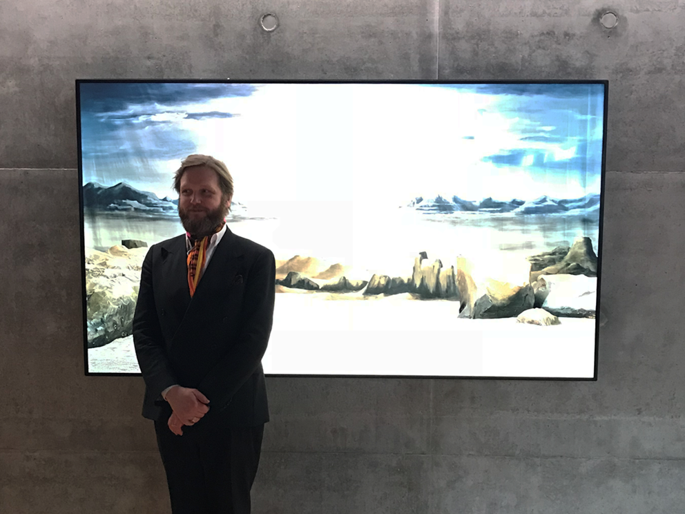 Den islandske kunstner Ragnar Kjartansson iklædt mørkt jakkesæt og slips foran skærmen med sit kunstværk, der hænger på en rå betonvæg