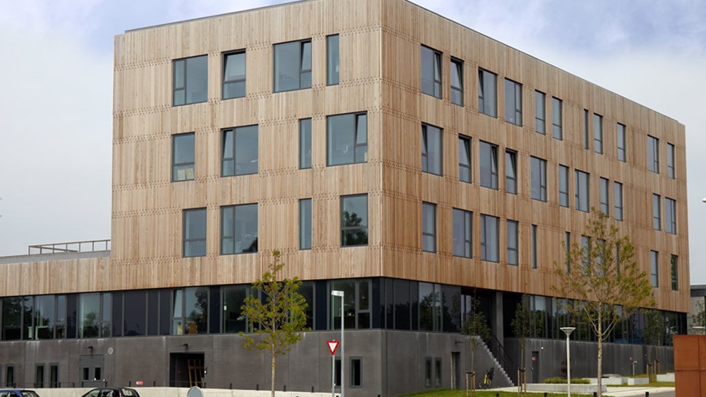 AAU Esbjerg. Bygningen har høj grå sokkel, hvorefter der er en etage med vinduesparti hele vejen, hvorefter bygningens tre etager er beklædt med trælameller, der brydes af store vinduer