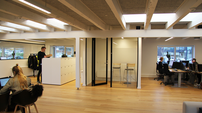 Bygningsstyrelsens kontor i Skanderborg. Medarbejdere sidder og arbejder ved et langbord og ved almindelige skriveborde. I baggrunden ses en medarbejder ved de halvhøje skabe i garderoben