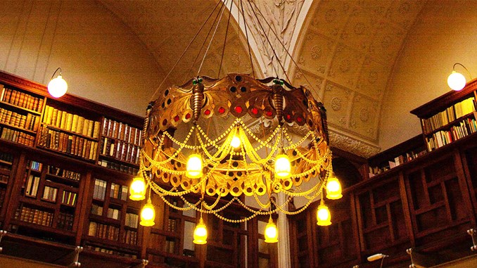 Den store lysekrone er tændt i biblioteksrummet med store hvælvinger i loftet fuldt ornamenteret af stuk, samt høje reoler i mørkt træ fulde af gamle bøger.